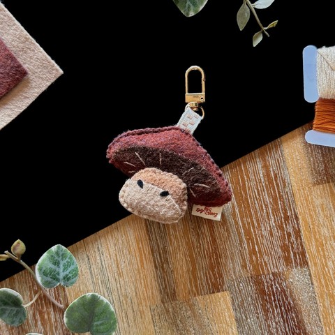 Picture of a handmade felt mushroom-shaped bag charm photographed on a black and wooden background; a few decorative plants and matching felt and threads around it. 
The bag charm is a brown mushroom, the cap is brown and angled backwards, revealing the bottom of it, made of a darker brown felt, a few lighter lines are embroidered on it. 
The foot is attached to the center of the underside of the cap, it is a two_toned beige, a darker one overcasting small suspicious eyes in the manner of a shadow. 
The bag charm is linked to a golden lobster clasp with a ivory ribbon checkered with brown.