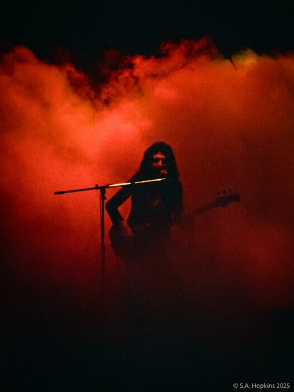 Color photo of John Deacon, bassist for Queen, playing on stage in a fiery red smokey blur.
