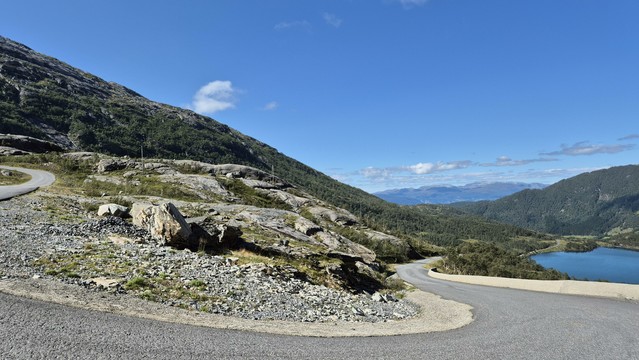 A U-turn in the Glacier Road up to Folgefonna above the timber line.
On the right there's a small lake fed by glacial water. Another mountain range can be seen in the back.
The road winds itself further up to the left between bare rocks and sparse vegetation.