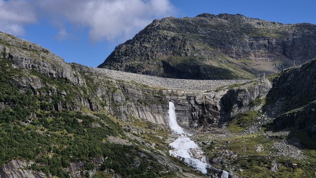 A waterfall pours out of a hole in a rocky cliff underneath a stone wall barrier against avalanches.