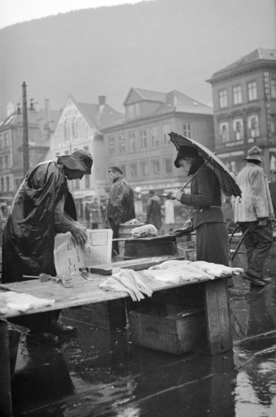 A black and white photo of a fishmonger in rain gear wrapping fish in a newspaper, likely selling it to the woman with an umbrella in front of him. In the background, a row of houses under a grey sky.