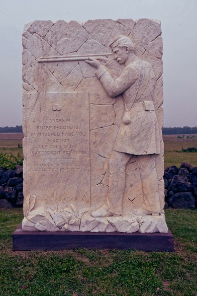A rectngular rock with a cracked pattern cut into it reminiscent of a stone wall with a bas relief of a rifeleman sighting in a gun and wearing a union army uniform of the civil war period in front of a stone wall in a great open field.
