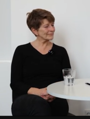 Eine Frau über 60, mit schwarzem Oberteil und dunkler Hose, sitzt mit übereinandergeschlagenen Beinen vor einer weißen Wand. Sie hat rötlicher Haar, vor ihr ein weißer Tisch auf dem ein Glas Wasser steht.