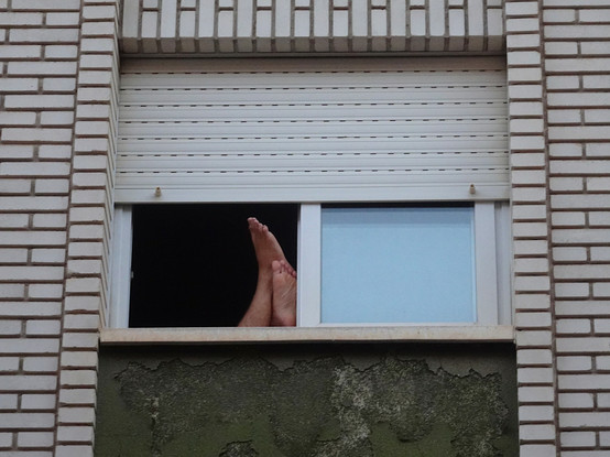 Plano cerrado de una ventana abierta, con la persiana a medio bajar, en la fachada de un edificio (ladrillos blancos). Apoyados en el alféizar y el marco se ven los pies de una persona.
