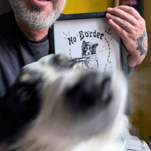 A bearded white man proudly showing a linocut with a No Border border collie.
In front of him, a border collie occupies the lower half of the picture. It's moving and blurry but we can you can see its nose ready to be booped