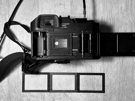 Monochrome processed photo of a rangefinder camera shown from the back with the film door open. In the film gate, a 3D-printed frame holds a focusing screen. Below the camera, three additional 3D-printed frames are laid out in a row, each fitted with a different material tested as a diffuser for focusing: tracing paper, Mylar foil, and a matte display protector.