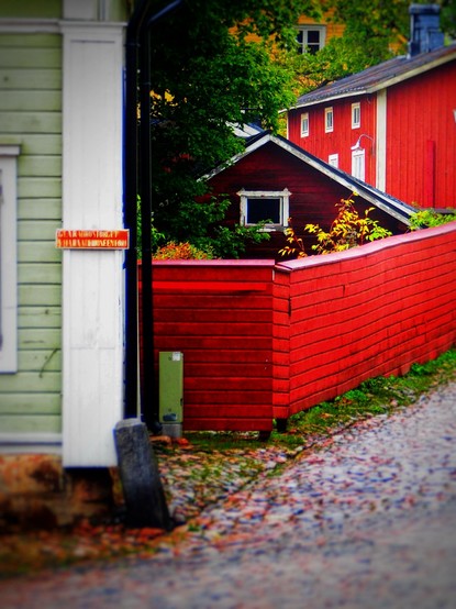 A narrow, cobblestone pathway leads through a quaint scene featuring colorful wooden houses of varying shades, predominantly red and green, with a fenced area and lush greenery. A signpost is visible on a building wall.