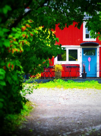 A vibrant red house is partially obscured by green foliage. The house features a blue door and a pink spoon decoration on the wall. In front, there are a few black chairs and a potted plant, with a cobblestone path.