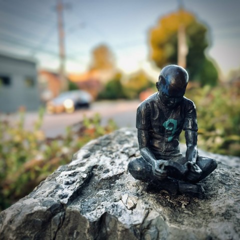A miniature sculpture, about six inches tall, of a young boy sitting and reading a book, rests on a large rock in the Ladd's Addition neighborhood on a sunny morning.