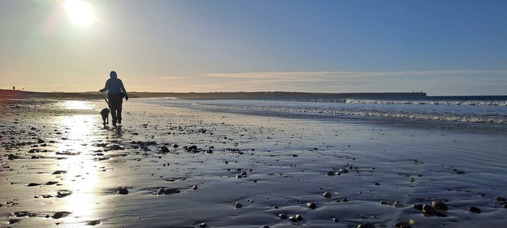 A long shot in landscape of me and Quintin (17 week old black labrador pup) walking away on a wide sandy beach. A headland in the distance forms the horizon and the calm sea laps gently to the right of the image. A beam of sunlight extends down onto the beach where the wet sand reflects the nearly early morning sun.