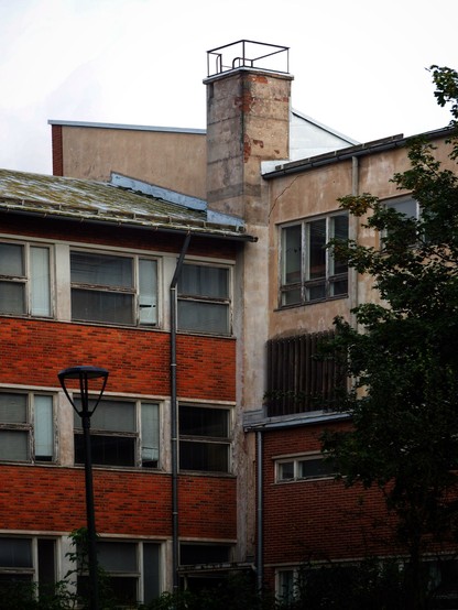 A building with a mix of brick and concrete walls. The structure features a tall chimney, several windows with a slightly aged appearance, and a lamppost in the foreground. Green foliage is visible near the base of the building.