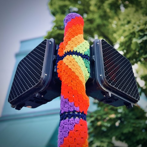What looks to be a homemade, crocheted rainbow-colored cover wraps around the single pole of two electronic walk signs on a street corner.