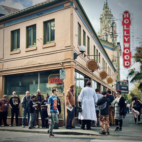 A human line forms around a building, with Hollywood Theatre and its vertical sign in the distance on a sunny day.