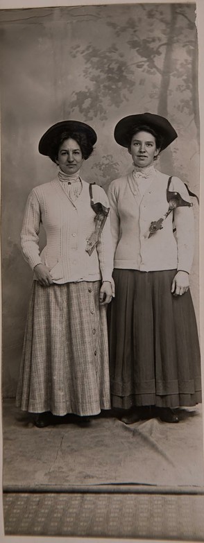 Two young women from 1910 dressed for ice skating. They are wearing long skirts, shirts with heavy sweaters and hats. Their ice skates are hanging by the straps over their shoulders. The photo looks to have been taken in a photo studio with a backdrop hanging down and covering part of the wood flooring.