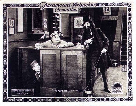 A poster for the film. Black and white. There are two men trying to hide behind a counter of a hotel front desk. There is a third man who is apparently threatening the other two. This third man is wearing a black hat, overcoat and trousers.