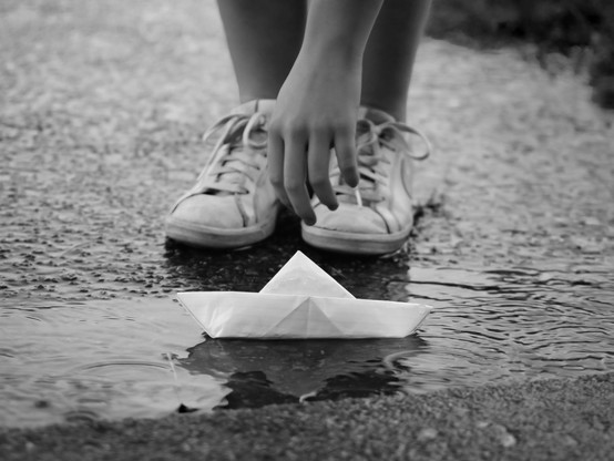 Un enfant se baisse, on voit sa main tendu qui essaye de ramasser un bateau en papier dans une flaque d'eau.