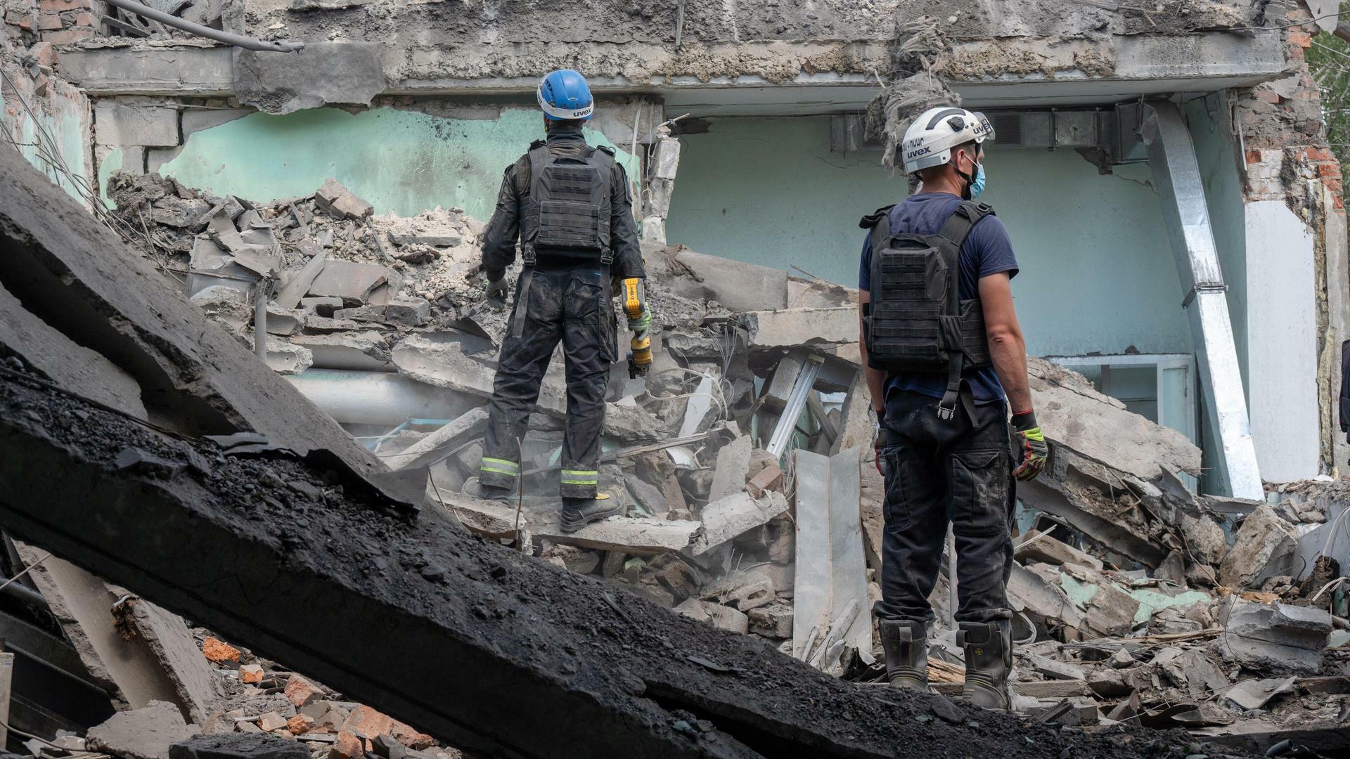 Ein zerstörtes Gebäude in der Ukraine. Auf den Trümmern stehen zwei Männer mit Schutzhelmen. 