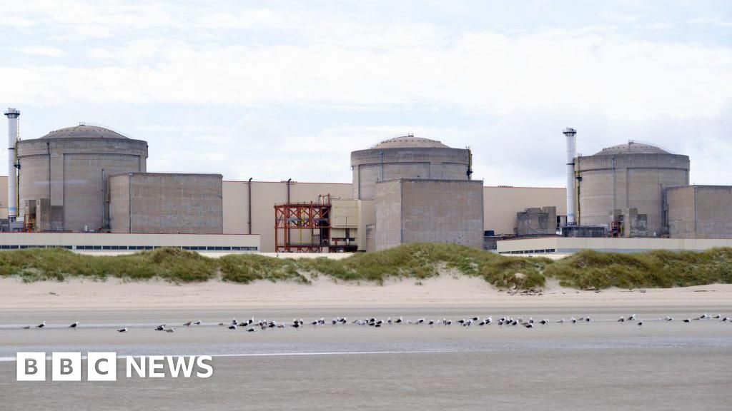 A still image of the Gravelines nuclear power plant, with three stone-coloured units connected by a large white wall, set behind a stretch of empty coast with the tide out.