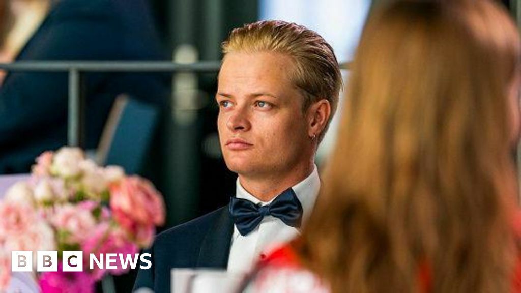 Marius Borg Høiby is wearing a black tuxedo and looking to his right while seated at a table. In the foreground, there are pink flowers on the table and a woman with her back to the camera.