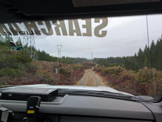 A dirt road running through a cleared strip of pine forest with high voltage transmission lines running to the left of it, seen through the windshield of a truck.

A radio microphone is hanging from a mount on the dashboard.