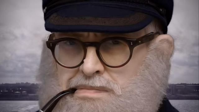 A tight close-up face of a young or middle-aged man dressed in a costume of a sea captain, or possibly lighthouse operator, sporting a dark hat that is part uniform and part cap, and holding a pipe (hand off screen).  Most prominently is a fake white beard that has a tidy but generous thickness all the way up to the ears, obscuring them.  The background is an obvious chroma-key of stock footage of the sea with a coastline a few minutes behind, trails showing departure.
