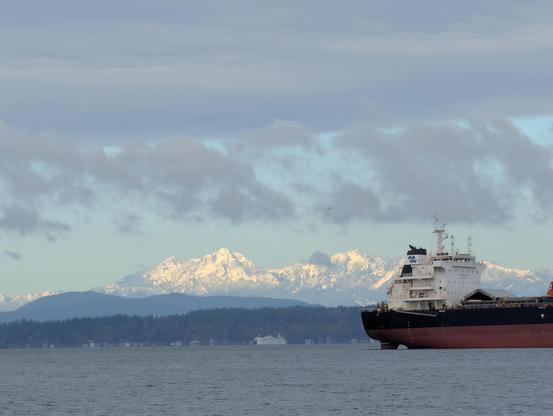 Line of snow capped mountains seen above lower foothills, seen across a large body of water. The stern of an anchored bulk cargo carrier is poking into the right side of the frame