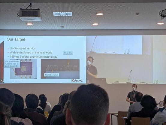 Me standing in front of a room full of conference goers, wearing a 3M Aura, presenting a slide showing a package overview and cross section of a 180nm 5 metal aluminum device