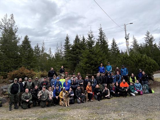 Group photo of several dozen people including a few sheriffs deputies, plus four dogs, in a forest clearing surrounded by tall evergreen trees
