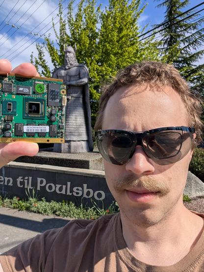Me, white male with light brown hair and a moustache wearing sunglasses, standing in front of a stone statue of a Viking with text "velkommen til Poulsbo" (Norwegian for "welcome to Poulsbo") on the base.

I'm holding a green PCB with ENIG finish prominently featuring a large flip chip BGA that has on die coupling caps and a weird L shaped gold stripe. This is the Intel "Poulsbo" southbridge