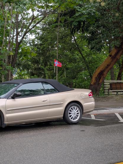 Parked car with a Norwegian flag on a wooden stick hovering in the air above the back of the cabin, presumably attached to a radio antenna too small and dark in color to be visible from this distance