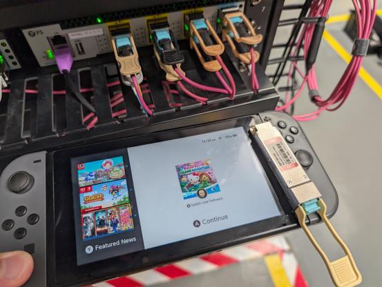Nintendo switch on the home screen (with Hello Kitty Island Adventure paused) with a 40Gbase-SR4 QSFP+ optic on top of it, held in front of a server rack with a 40/100G switch on it