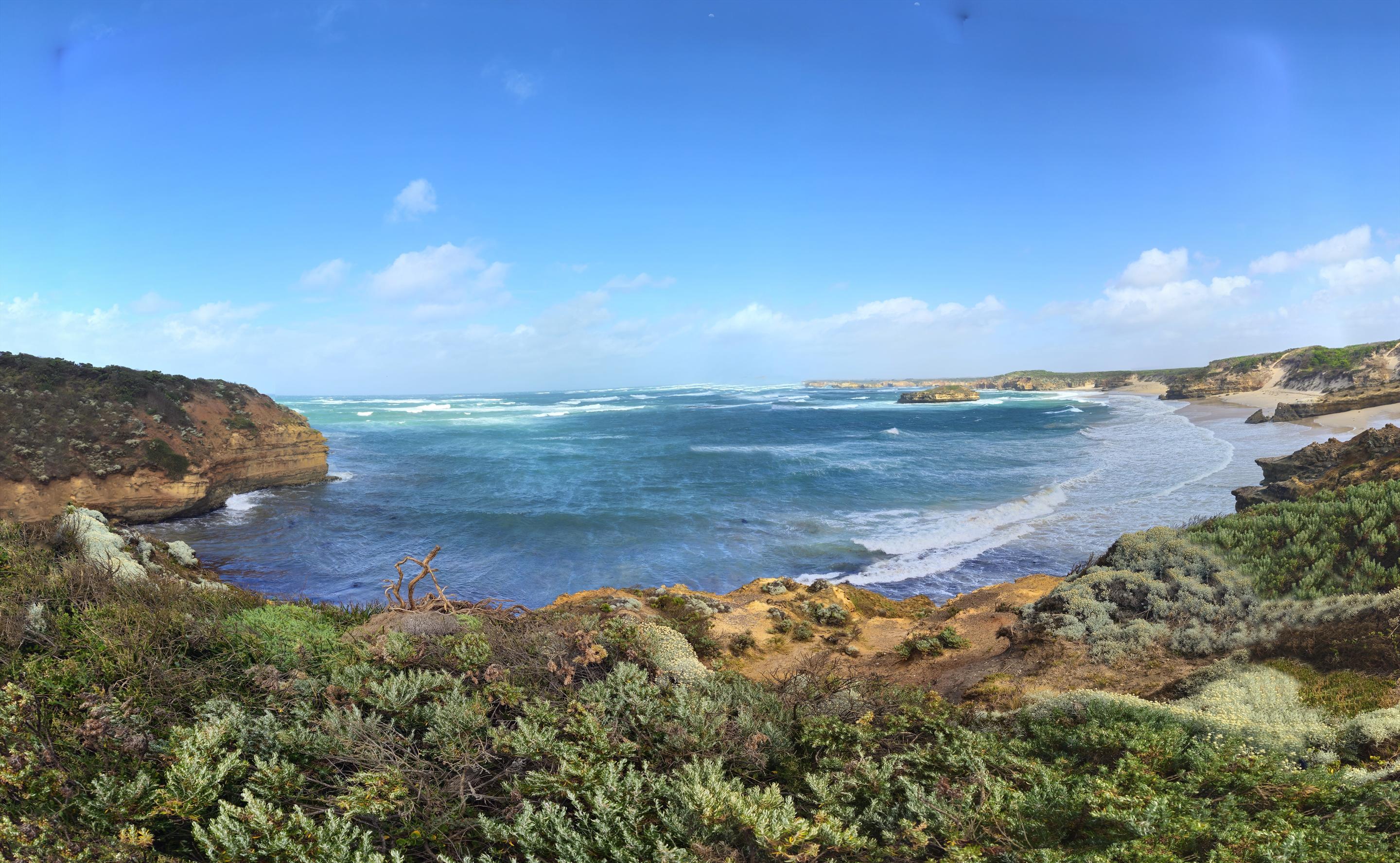 Wide panoramic view of the Bay of Martyrs on Australia's Great Ocean Road under a deep blue sky. Turquoise and dark blue ocean fills the frame, with lines of white breakers rolling into a curved sandy beach on the right. Golden limestone cliffs frame both sides of the bay, and a small flat-topped limestone island stack sits offshore. The foreground is dense low coastal scrub in silvery green, with a patch of weathered driftwood on the cliff edge. Distant headlands fade into haze along the horizon.

Bay of Martyrs is on the Great Ocean Road near Peterborough, Victoria, part of the Bay of Islands Coastal Park. The name (along with nearby Massacre Bay and Massacre Hill) is tied to local accounts that European settlers in the 1840s drove Girai wurrung (Kirrae Whurrong) people off these cliffs to their deaths during the violent dispossession of the Western District. There's no surviving written record of the specific event – which is itself part of the story