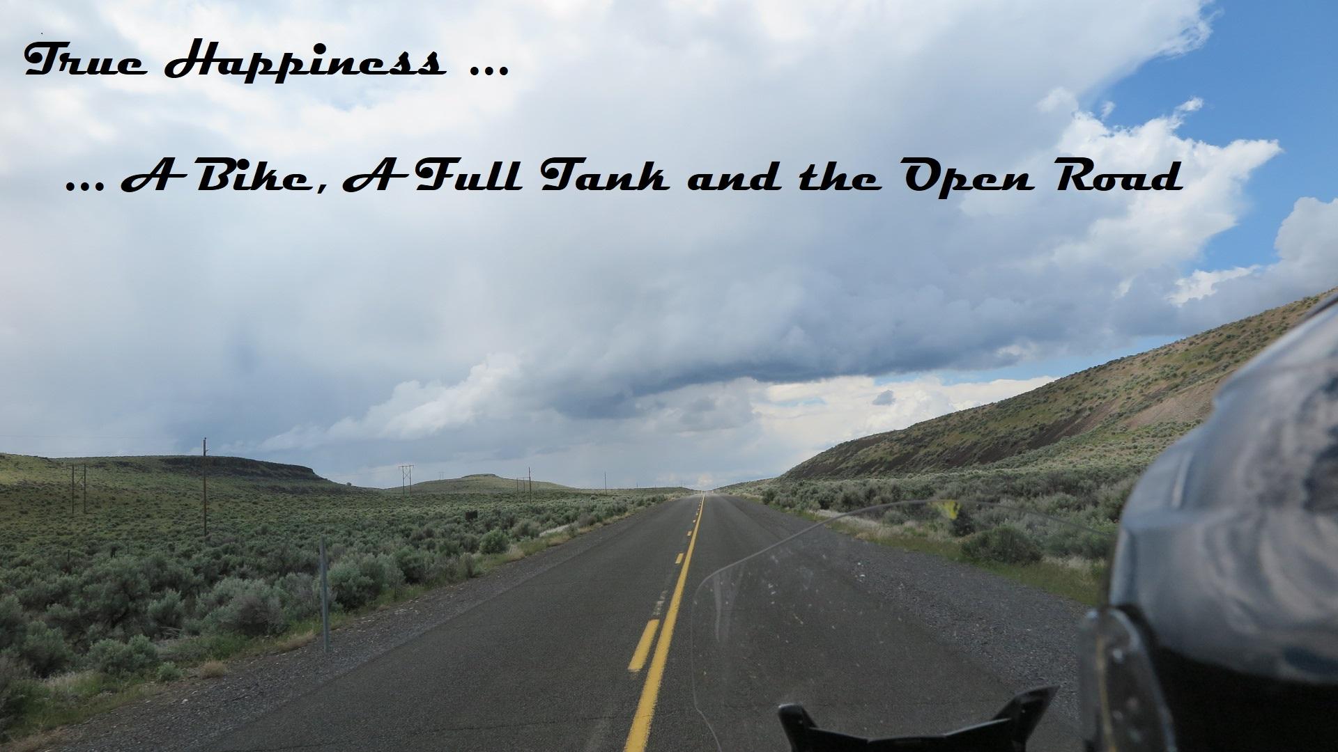 Rider's-eye view over a motorcycle windscreen. A two-lane asphalt road with a yellow centre line runs dead straight to a vanishing point on the horizon, through flat high-desert sagebrush. A low green-brown ridge with exposed rock rises on the right; power poles march away on the left. Ahead, a large grey storm cloud spreads across an otherwise bright blue sky, with rain visible falling in the distance. The bike's dark fairing and mirror occupy the lower right corner. Overlaid text in black script reads: "True Happiness ... A Bike, A Full Tank and the Open Road."
