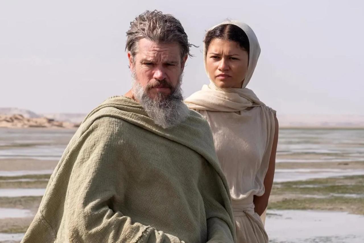 Matt Damon, with gray beard and in coarse beige robes, standing in front of Zendaya, with off white robes and shawl revealing her hair parted down the middle and pulled back into [implied] side buns. They are standing in front of sand and rocks, though on closer inspection there is also a gray sea under a hazy sky. They are illuminated by [implied] two bright suns