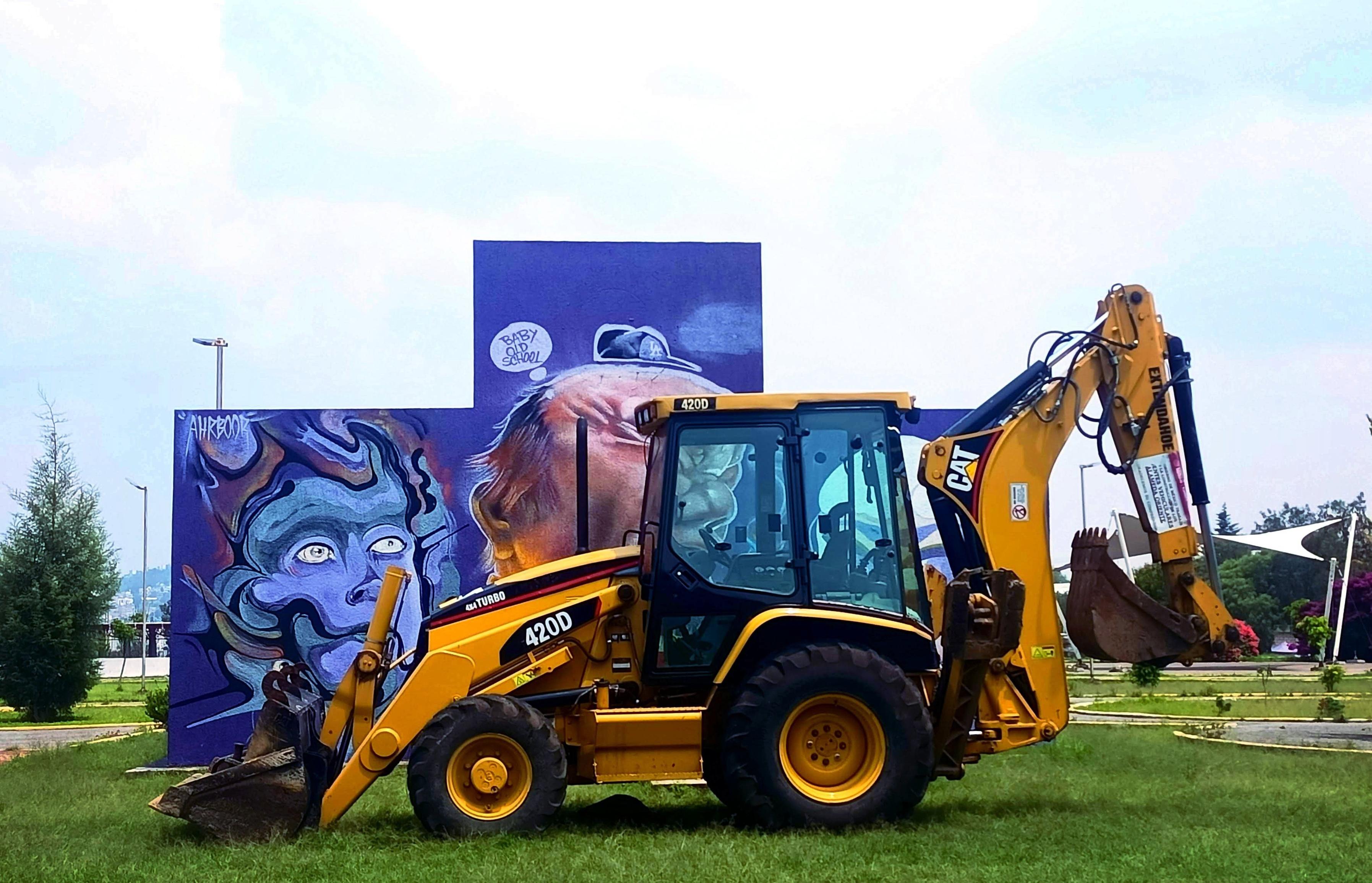 a photo of an excavator in front of an art wall in a park, where the workers come to play soccer.