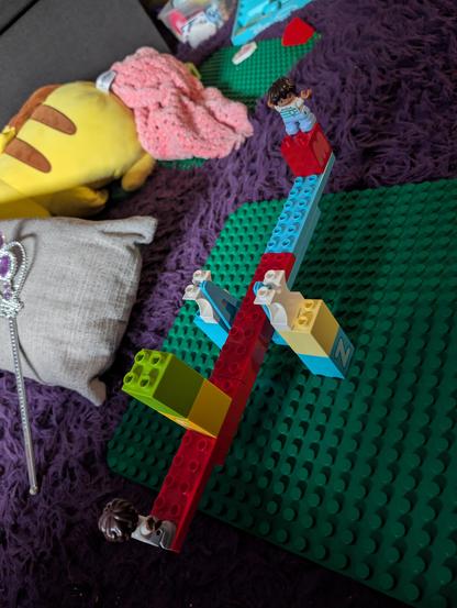 Lego balance beam with two masses, one at the end of the arm and the other at the midpoint but twice the size, still balanced.
A silver magic wand with purple gems is on a pillow in the background