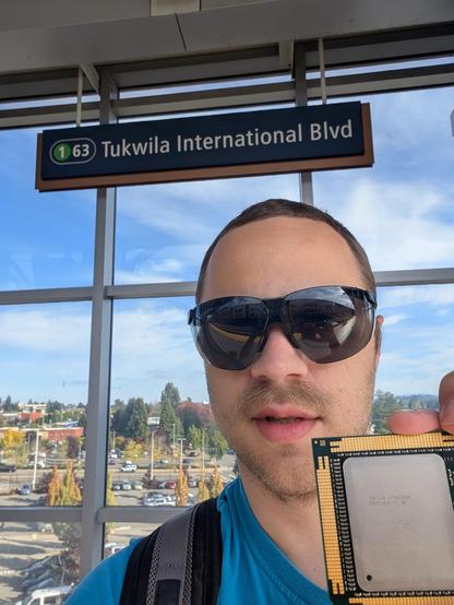 Selfie of me, white male with sunglasses and some stubble, holding a large Intel CPU package in front of the Tukwila International Blvd light rail station