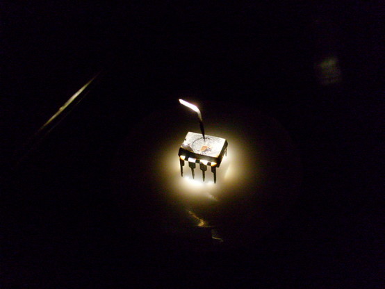 Decapsulated IC in an 8-pin DIP package sitting on a microscope stage in a darkened room, with the light from the objective shining down on the chip in a manner reminiscent of some kind of torture chamber or interrogation room.

A microprobe needle hovers menacingly just above the silicon surface.