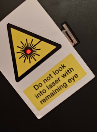 The Red LED of a Thinkpad Lid shining out of the middle of a triangular laser warning sign sticker. Text below it reads:"Do not look into laser with  remaining eye"