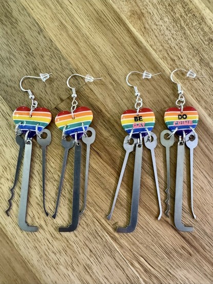 Two pairs of functional lockpick earrings. The heart-shaped charm is hand-made from UV coated polymer clay displaying the colors of the Pride flag. One pair has lettering on top of the flag colors that says "Be Gay" and "Do Crime". The lockpicks profiles were designed by the notable Christina Palmer.
