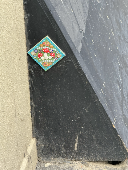 A brightly decorated ceramic tile featuring three bejewelled Amanita Muscaria stuck on the side of a temporary street fixture 