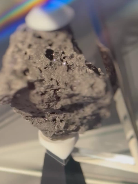 A video with tight zoom and shallow depth of field, tracking around an exhibit of a piece of moon rock, glistening at certain angles.  There are reflections from the protective glass case, and the rock is pockmarked with holes, fastened with white plastic handles above and beneath