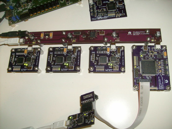 Closeup of one board showing a very long, skinny USB hub made on a bare purple PCB with a barrel jack and mini-B jack along the west side and regularly spaced male mini-B ports along the south edge, each connected to a devkit of some sort