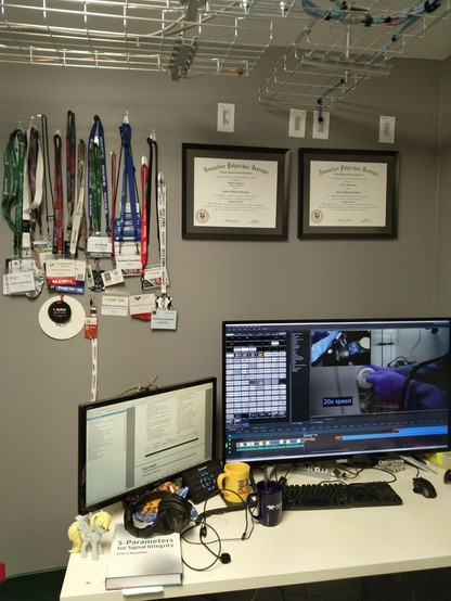 Photo of my desk. A PDF of a chip datasheet is open on a 24" monitor to the left while kdenlive is open on a 43" display to the right, editing a video of me working inside a fume hood.

Mugs with Thorlabs and Teledyne LeCroy logos sit to the left of the keyboard next to a headaet and a copy of "S-Parameters for Signal Integrity" and a Derpy Hooves pony toy.

Hanging up on the wall above the computer screens are a large ensemble of conference badges and framed copies of my BS and Ph.D degrees.