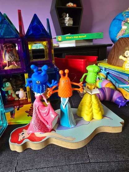 Three Disney princess toys with rubber alien finger puppets over their heads.

Princess Elsa has an orange monster with four arms and two eye stalks instead of a head.

Belle has a more humanoid green one.

Aurora (I think) has a blue critter with antennae, one eye, and covered in spikes.