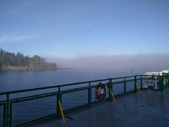 View from the top deck of a large boat looking over a green railing with an orange life ring mounted to it.

The sky is clear blue with very few clouds but a dense fog bank obscures the water and shoreline to the right of the frame