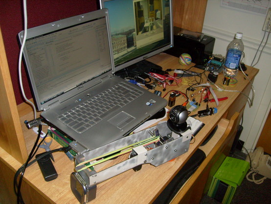 Sheet metal and aluminum rubber band launcher with a webcam mounted to the top of it sitting on a desk next to a laptop, external monitor, and a pile of assorted embedded development tooling.

The laptop is propped up on a copy of "The Personal Computer from the Inside Out" to make its internal screen more closely match the height of an external monitor sitting next to it.