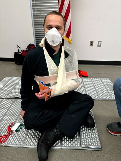 Me, mid 30s white male, wearing a N95 and black sweater and rain pants. I'm sitting on a foam pad on a concrete floor, with my arm firmly secured by a pair of orange SAM splints, various athletic wraps, and a sling fashioned out of a triangular bandage.