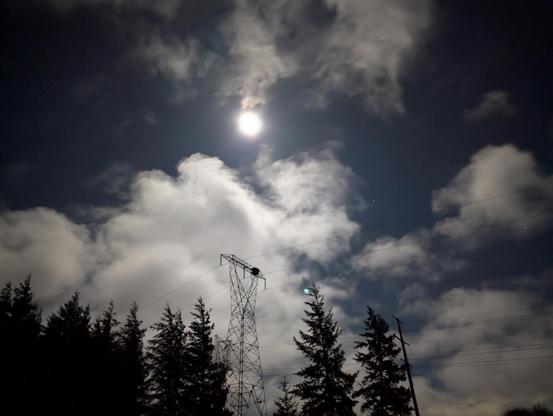 Full moon lighting up a cloudy sky at night. A line of tall evergreens form the horizon to the left, with a tall 3-phase transmission tower silhouetted against the sky above the trees