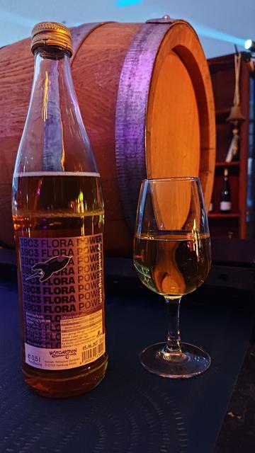 Bild mit Flora Mate Flasche und halb vollem Sektglas auf dem Tresen der Winzerbar. Im Hintergrund ein Holzfass und ein Regal mit Weinflaschen